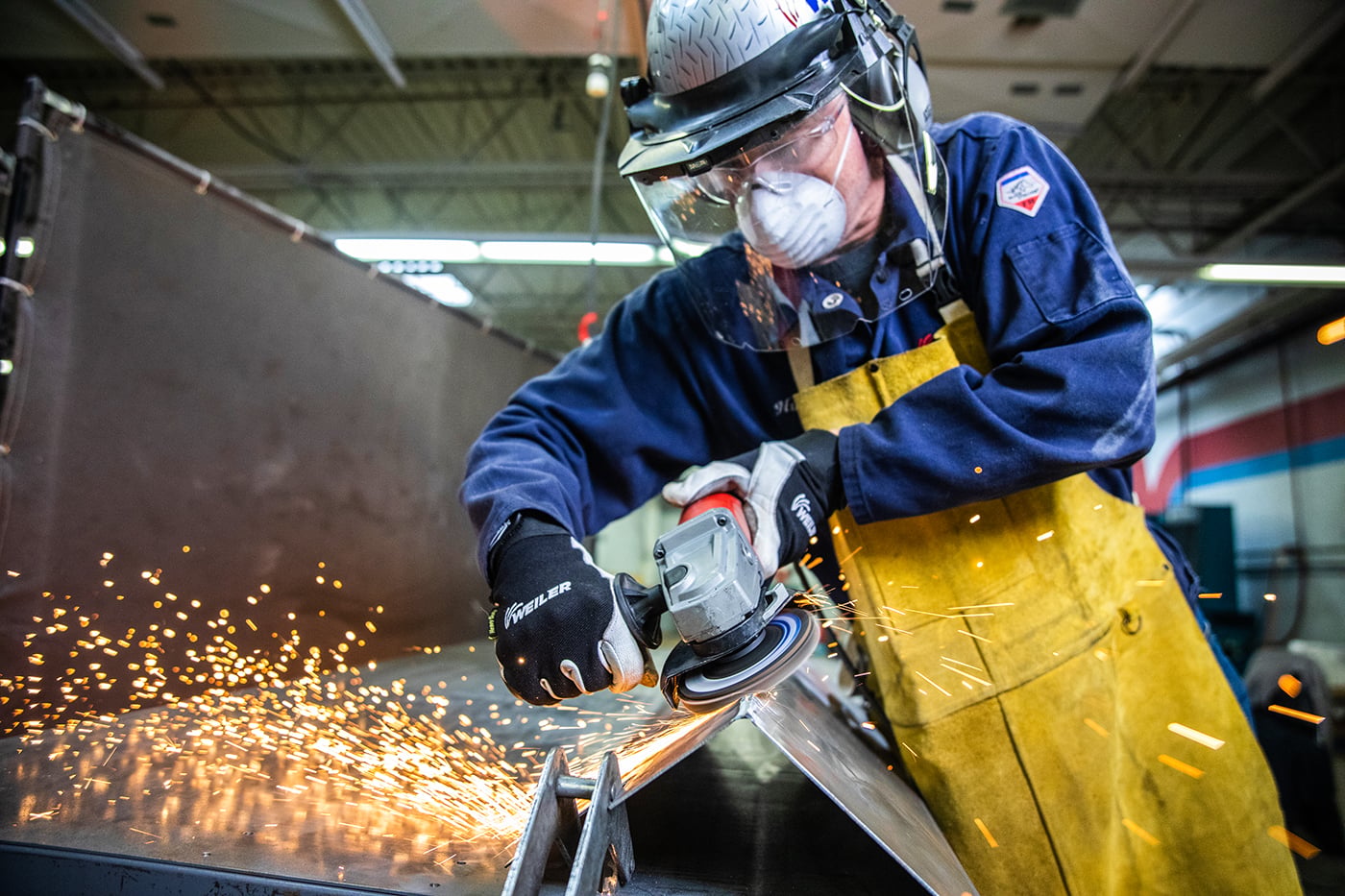 Metalworker weld grinding an angled workpiece and wearing full protective gear.